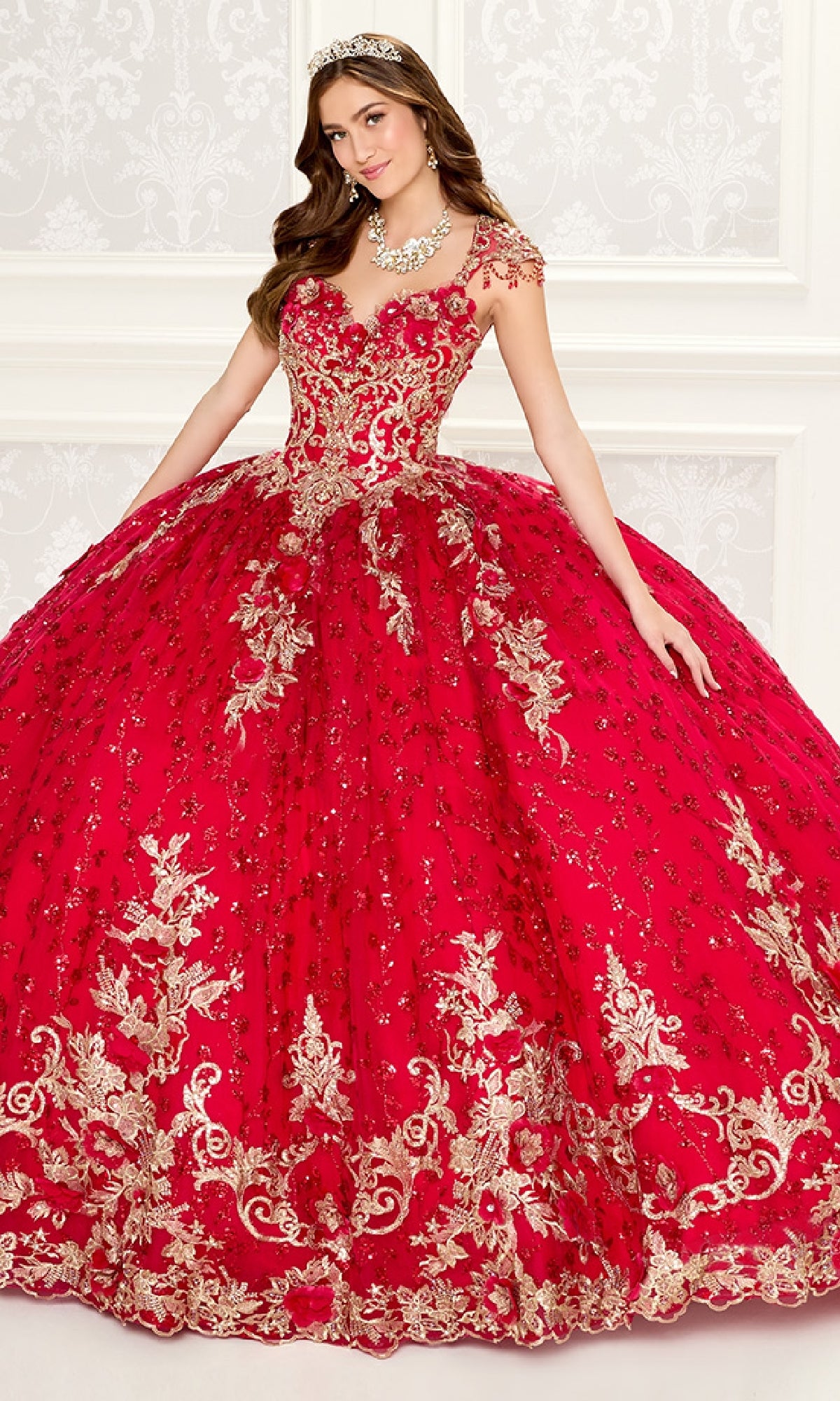 A woman in the Quinceanera Dress PR30088 By Princesa, featuring bright red fabric and gold embroidery, wears a tiara while standing before an ornate white-patterned backdrop.